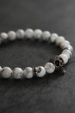 The Relentless Betrayal Until Death Storm Cloud Bead Bracelet, featuring white marbled beads and a metallic skull charm, is displayed on a dark, textured surface.