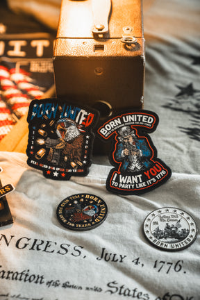 Assorted patriotic patches and pins with American flags and eagles from Born United Private Label are displayed on historic-text fabric. The Born United Patriot Box is in the background, commemorating America’s 250th Birthday.