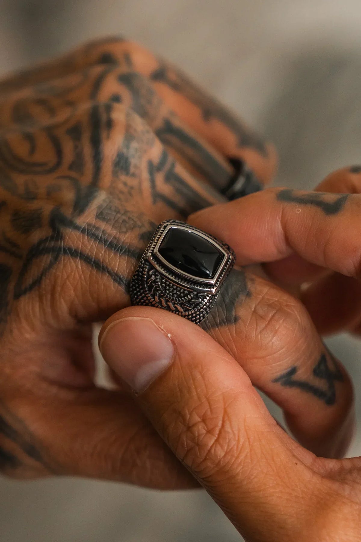 A tattooed hand wears The Aztec Ring by Relentless Betrayal, an ornate handcrafted piece featuring a large rectangular black stone, while another hand adjusts it.