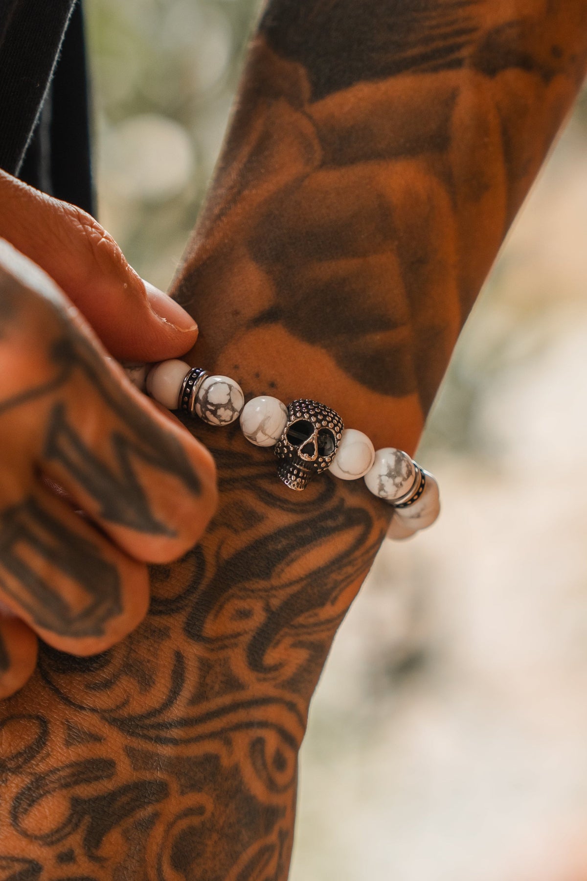 A tattooed hand wears the Relentless Betrayal Until Death Storm Cloud Bead Bracelet, featuring a decorative skull centerpiece and white marbled beads, shown in close-up detail.