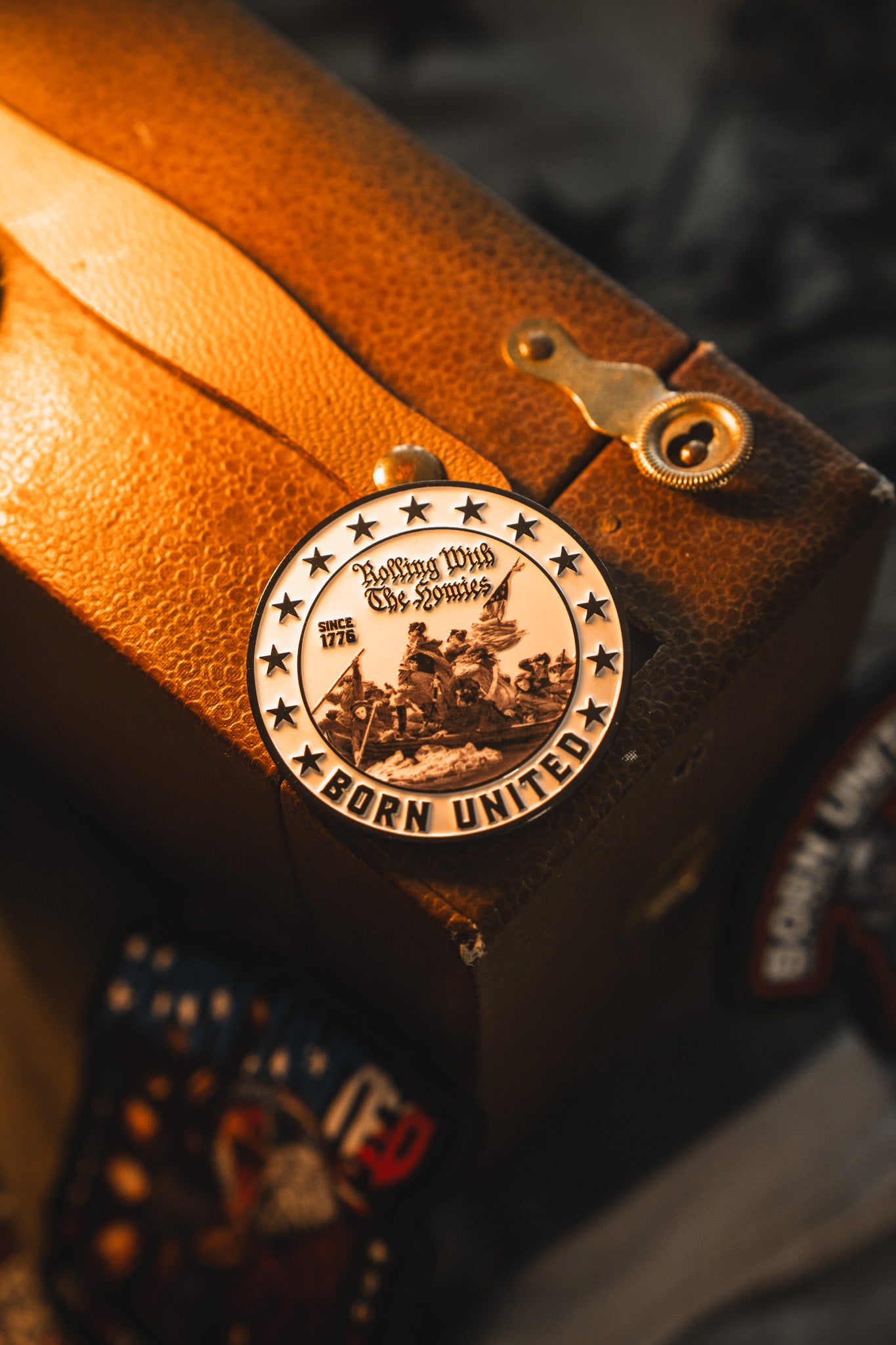 The Born United Patriot Box by Born United Private Label features a round metal medallion with stars, “Born United,” and “Rolling With The Syndicate Since 1776,” celebrating America’s 250th birthday atop a brown leather suitcase.