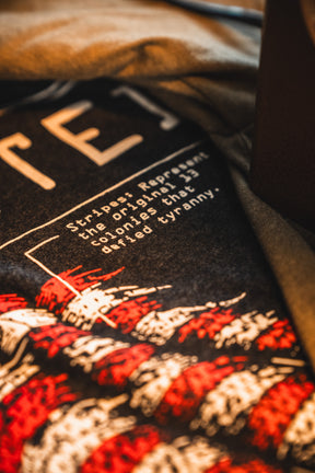 Close-up of a Born United Patriot Box Limited Edition Tee by Born United Private Label, showcasing “Stripes represent the original 13 colonies that defied tyranny” text and bold red and white stripes for America's 250th Birthday.