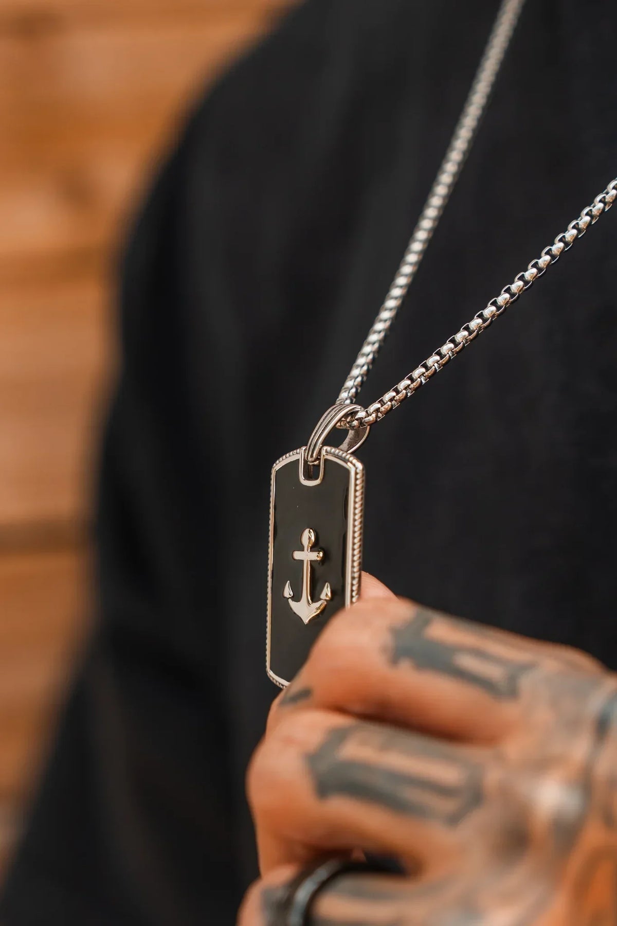 A tattooed hand holds the Relentless Betrayal Captain’s Curse Necklace, showcasing its handcrafted rectangular pendant adorned with an anchor symbol; the person is dressed in a black shirt.