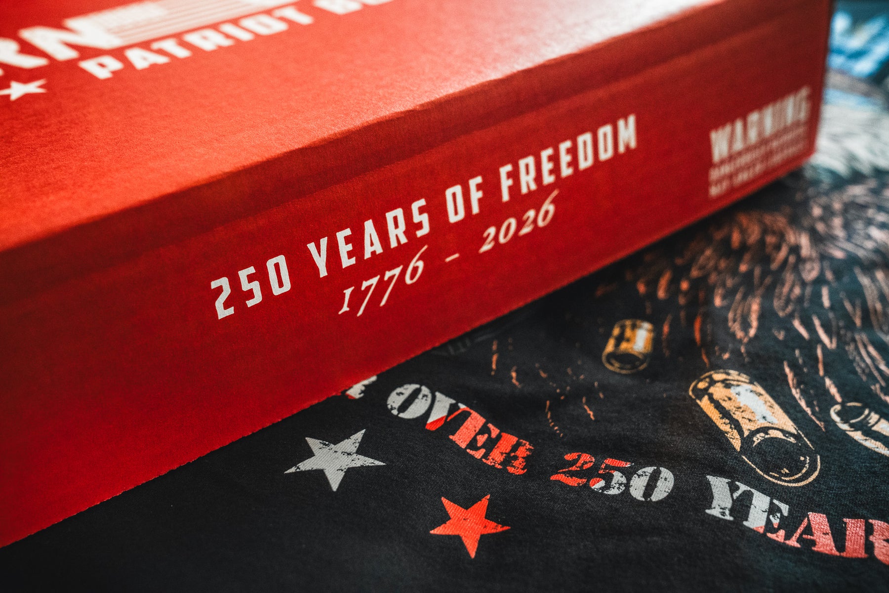 Red box with white text reading 250 Years of Freedom 1776–2026 and a partial Warning; the Born United Patriot Box from Born United Private Label rests on a dark surface with bullet and star images, celebrating America's 250th Birthday.
