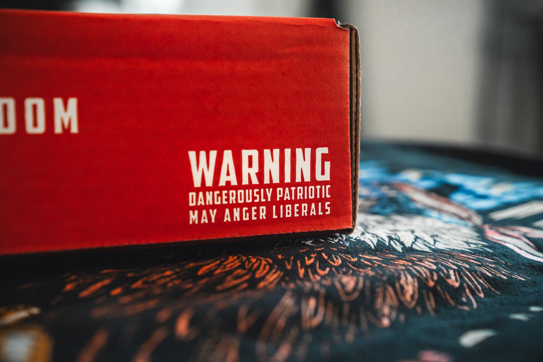 A close-up of the red Born United Patriot Box by Born United Private Label, featuring bold white text: WARNING Dangerously Patriotic May Anger Liberals. Part of an eagle illustration appears in the background—perfect for America's 250th Birthday limited tees.