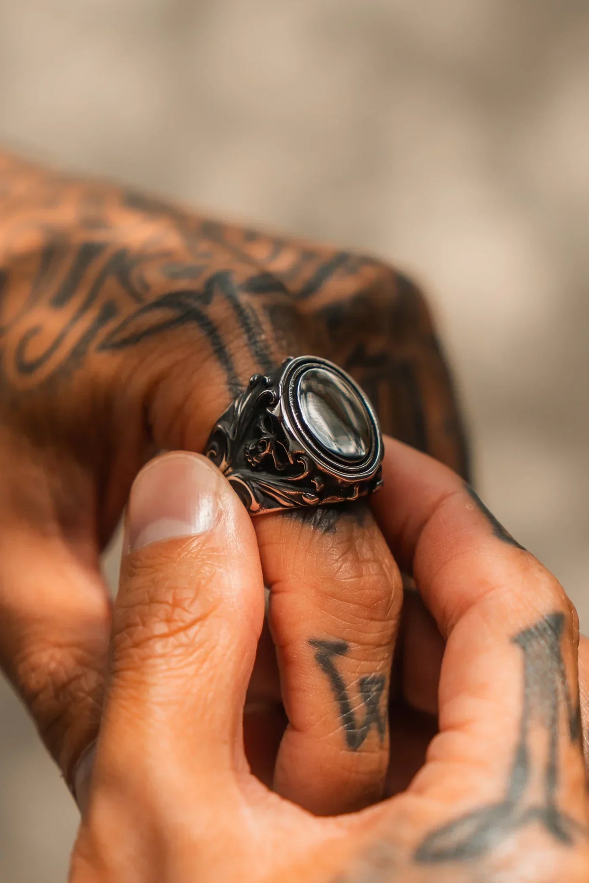 A tattooed hand holds the Relentless Betrayal No Face Ring, featuring a dark, polished stone and intricate metalwork, set against a blurred background.