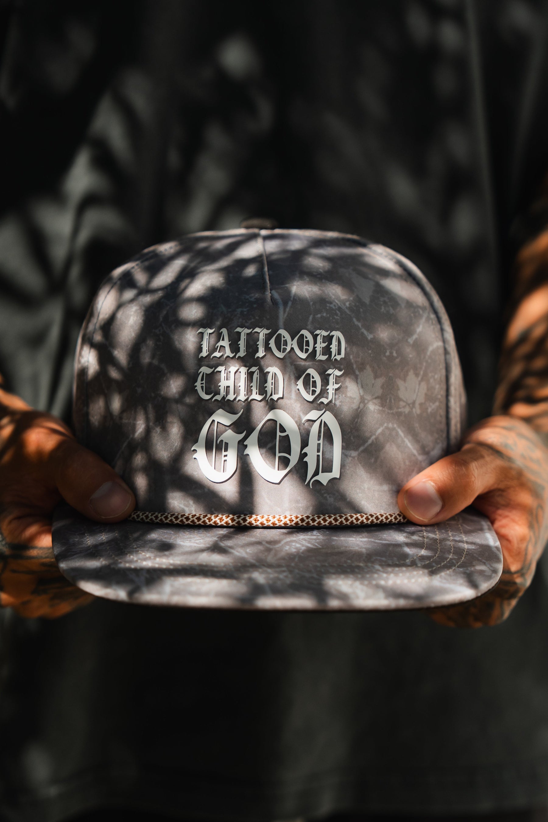 A person in a black shirt holds the Relentless Betrayal Tattooed Child of God Deadwood Camo PVC Snapback, its white gothic lettering and leaf shadows highlighting this bold faith-inspired accessory.