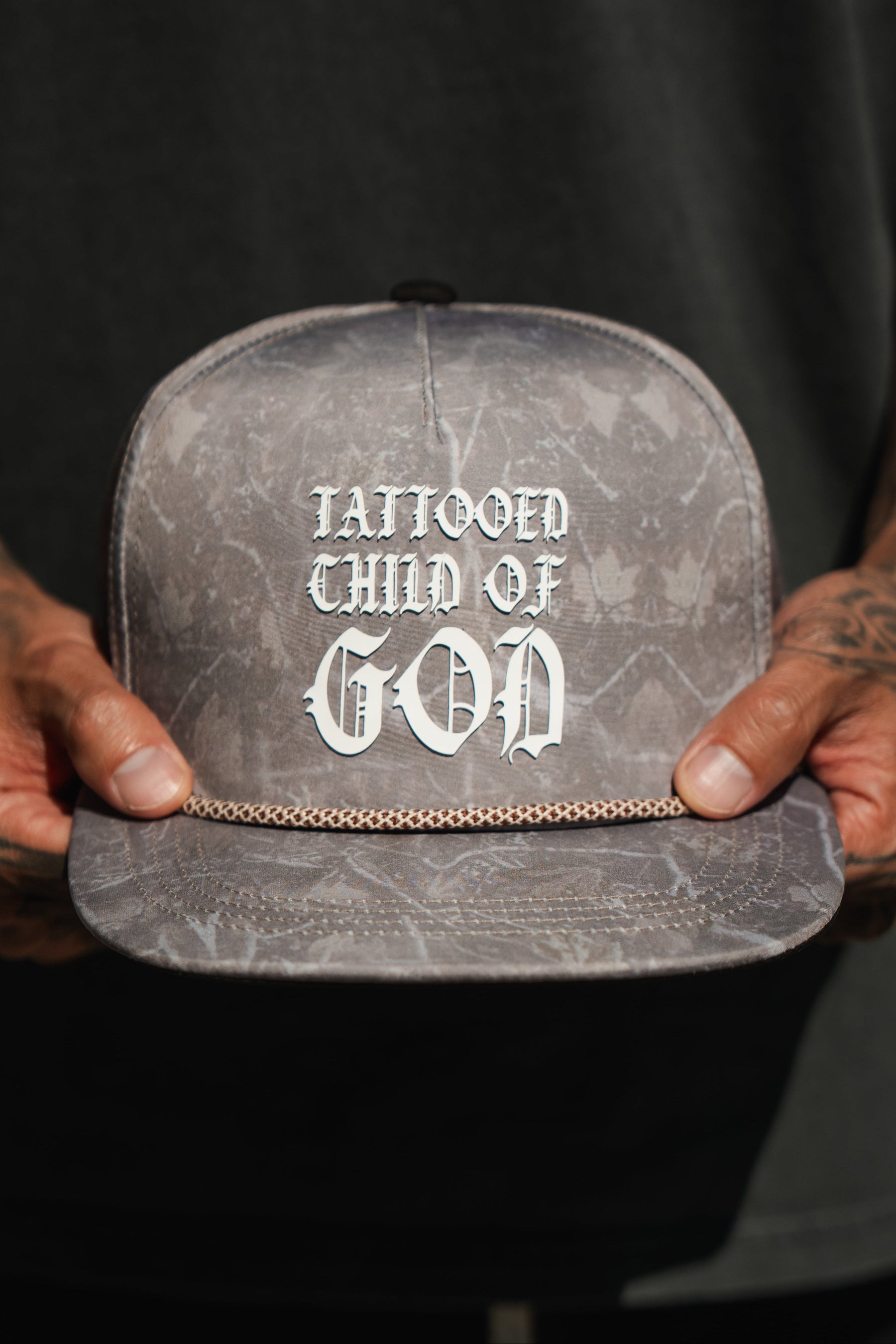 A person with tattooed hands holds a Tattooed Child of God Deadwood Camo PVC Snapback by Relentless Betrayal—a gray Christian cap with bold white text and a small gold chain across the brim.