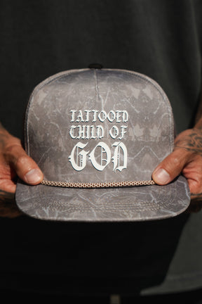 A person with tattooed hands holds a Tattooed Child of God Deadwood Camo PVC Snapback by Relentless Betrayal—a gray Christian cap with bold white text and a small gold chain across the brim.