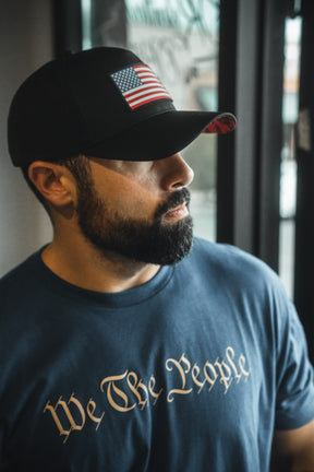 Man wearing a cap with an American flag and a 'We The People' t-shirt indoors.