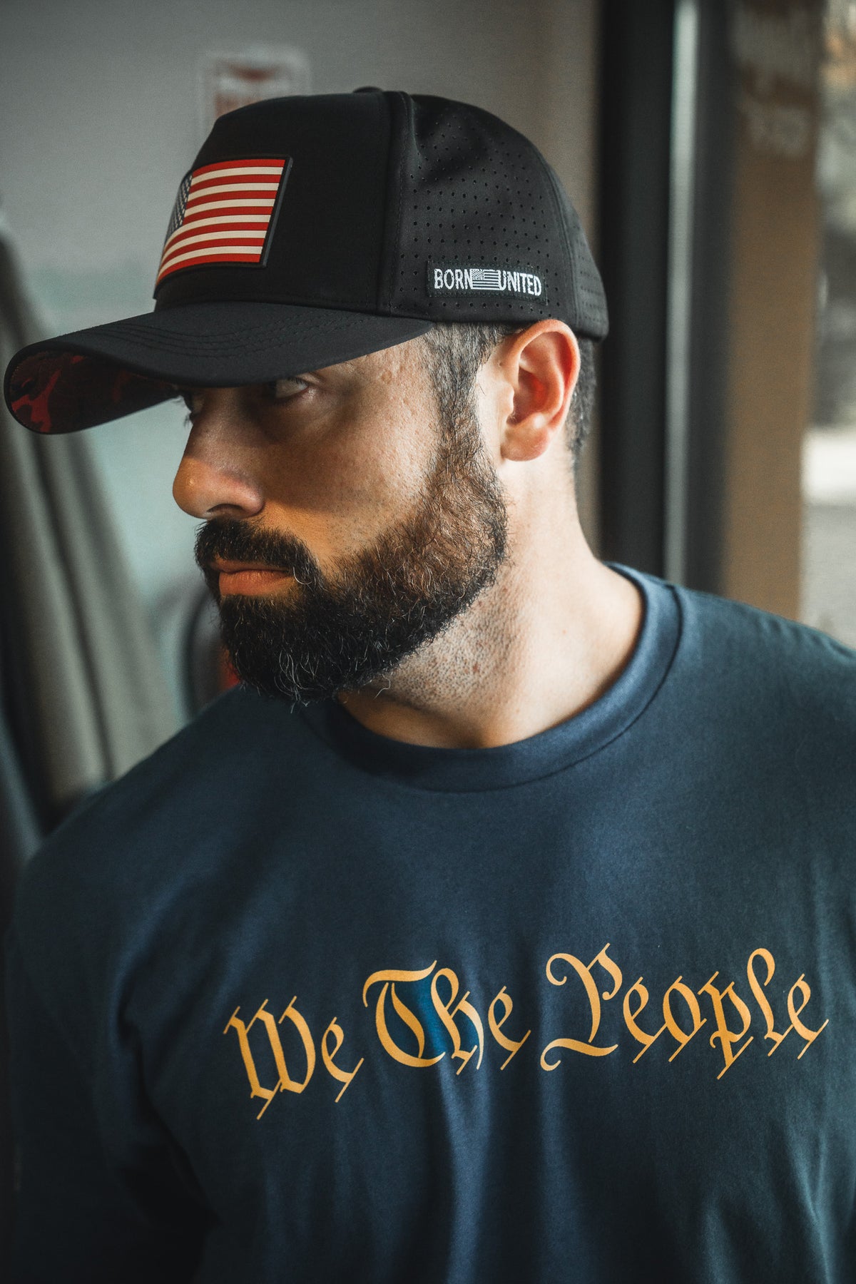 A bearded man in a black cap with an American flag patch and a blue "We the People" shirt by Born United Private Label looks to the side indoors, embodying unity and American pride.