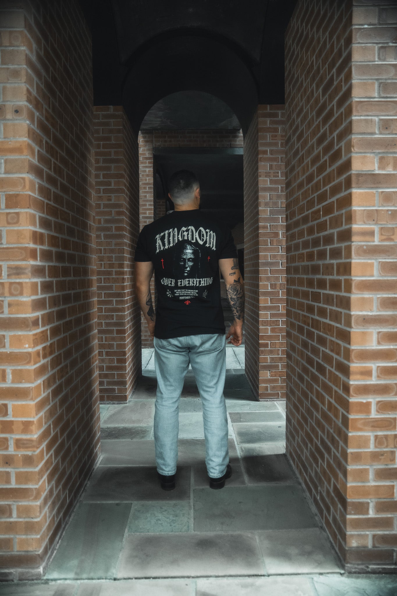 Wearing the Kingdom Over Everything Premium Tee by 13th and light blue jeans, a tattooed person stands in a brick corridor with arches, framed by tall pillars—reflecting on seeking God's Kingdom and His righteousness.