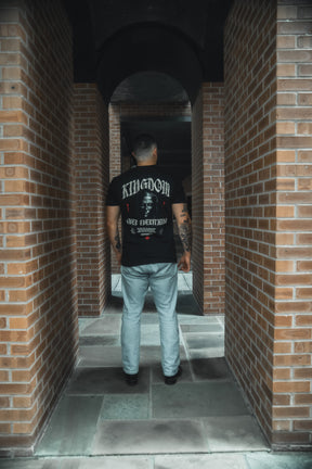 Wearing the Kingdom Over Everything Premium Tee by 13th and light blue jeans, a tattooed person stands in a brick corridor with arches, framed by tall pillars—reflecting on seeking God's Kingdom and His righteousness.