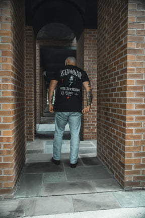 A tattooed person faces an arched brick hallway, wearing 13th’s Kingdom Over Everything Premium Tee—black, with “Matthew 6:33” and “Kingdom of God” printed on the back.