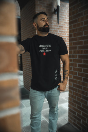 A bearded man stands by a brick wall, wearing the 13th Kingdom Over Everything Premium Tee featuring “Matthew 6:33” and “His righteousness” in white and red text, paired with light blue jeans. He looks to the side with one hand in his pocket.