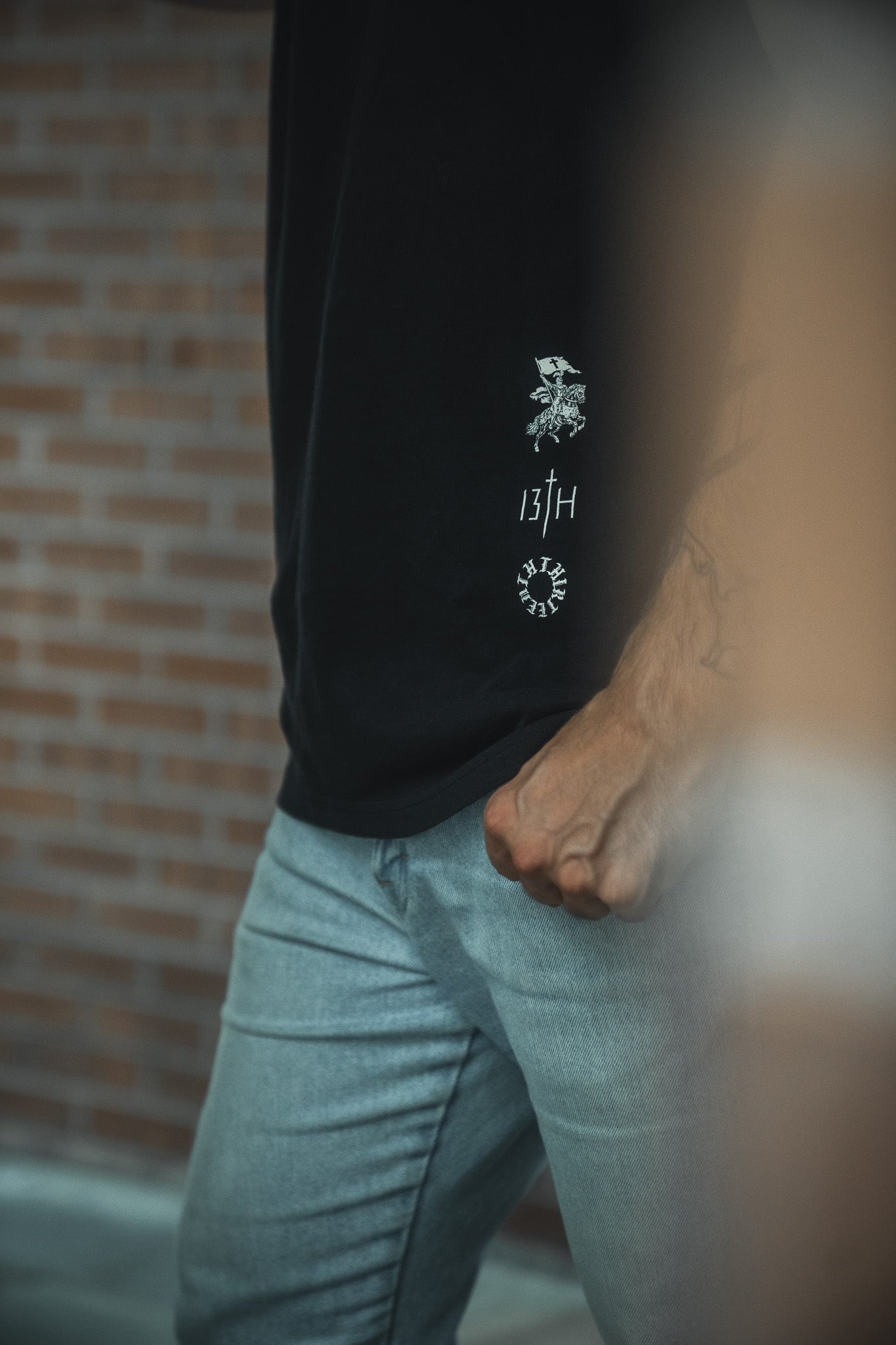Someone stands near a brick wall wearing light gray jeans and the Kingdom Over Everything Premium Tee by 13th, a black shirt with white graphics featuring "13TH" and a Matthew 6:33 reference. Their bent arm appears partially in the foreground.