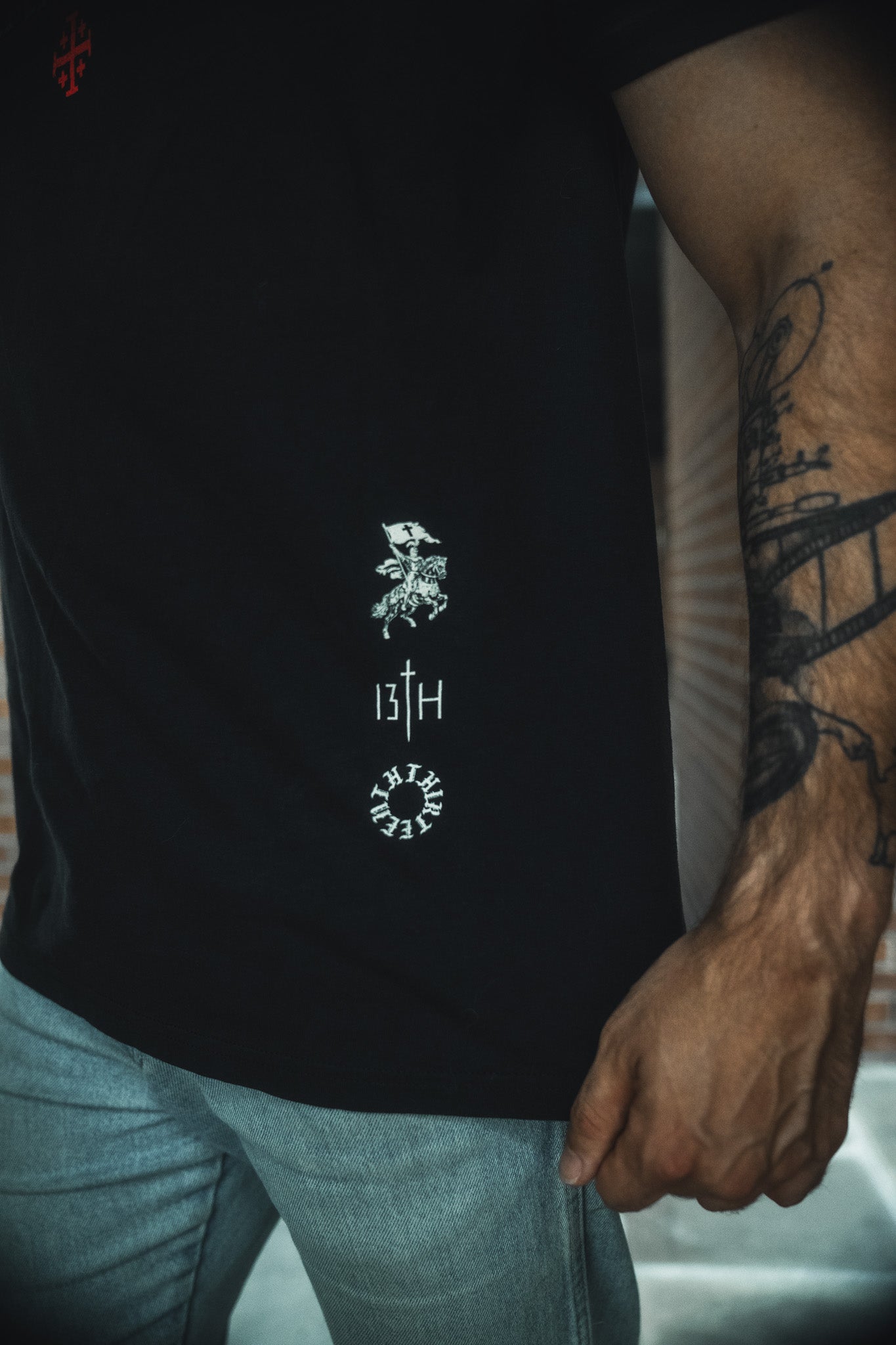 A person wears the 13th Kingdom Over Everything Premium Tee—black with white graphics and "13TH" text on the side—with light blue jeans. Their tattooed arm, partially visible, subtly references the Kingdom of God.