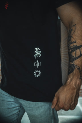 A person wears the 13th Kingdom Over Everything Premium Tee—black with white graphics and "13TH" text on the side—with light blue jeans. Their tattooed arm, partially visible, subtly references the Kingdom of God.