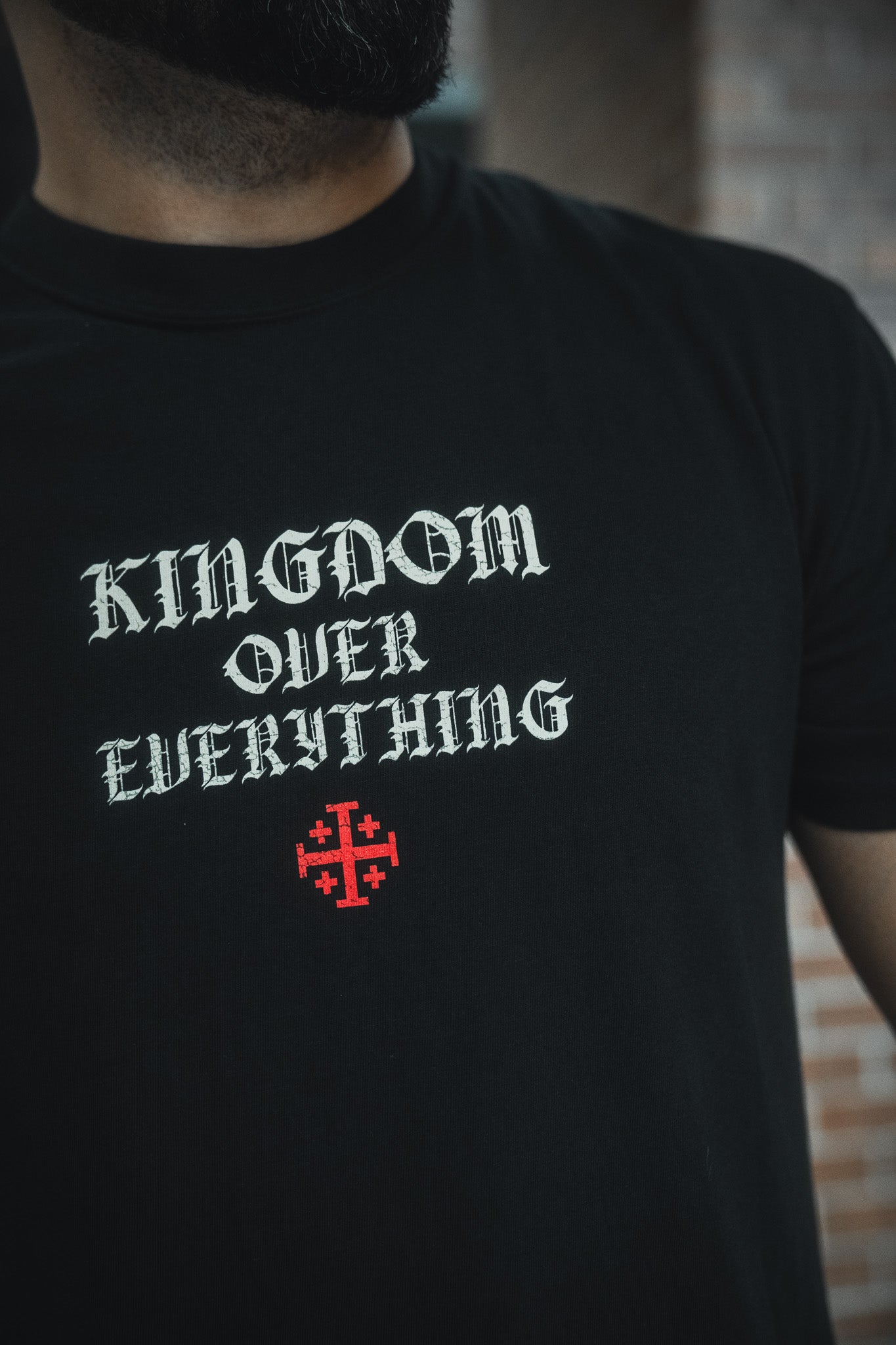 Someone wears the 13th Kingdom Over Everything Premium Tee—a black shirt with KINGDOM OVER EVERYTHING in bold white gothic font, a red cross below, inspired by Matthew 6:33 to remind us to seek first God’s Kingdom.