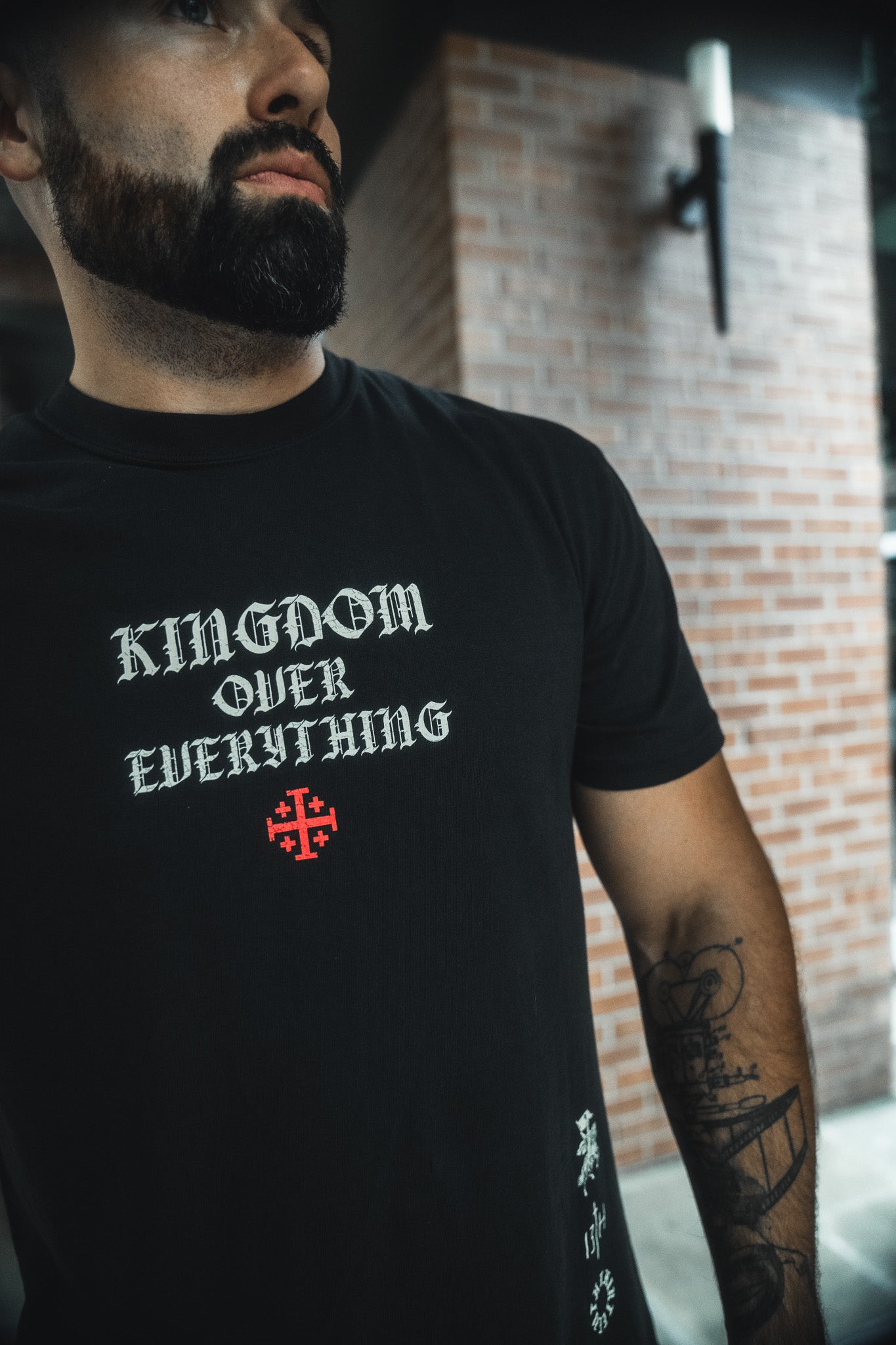 A bearded man with a tattoo on his left forearm stands before a brick wall, wearing the 13th Kingdom Over Everything Premium Tee featuring "KINGDOM OVER EVERYTHING" and a red cross beneath, inspired by Matthew 6:33.