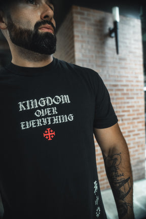 A bearded man with a tattoo on his left forearm stands before a brick wall, wearing the 13th Kingdom Over Everything Premium Tee featuring "KINGDOM OVER EVERYTHING" and a red cross beneath, inspired by Matthew 6:33.