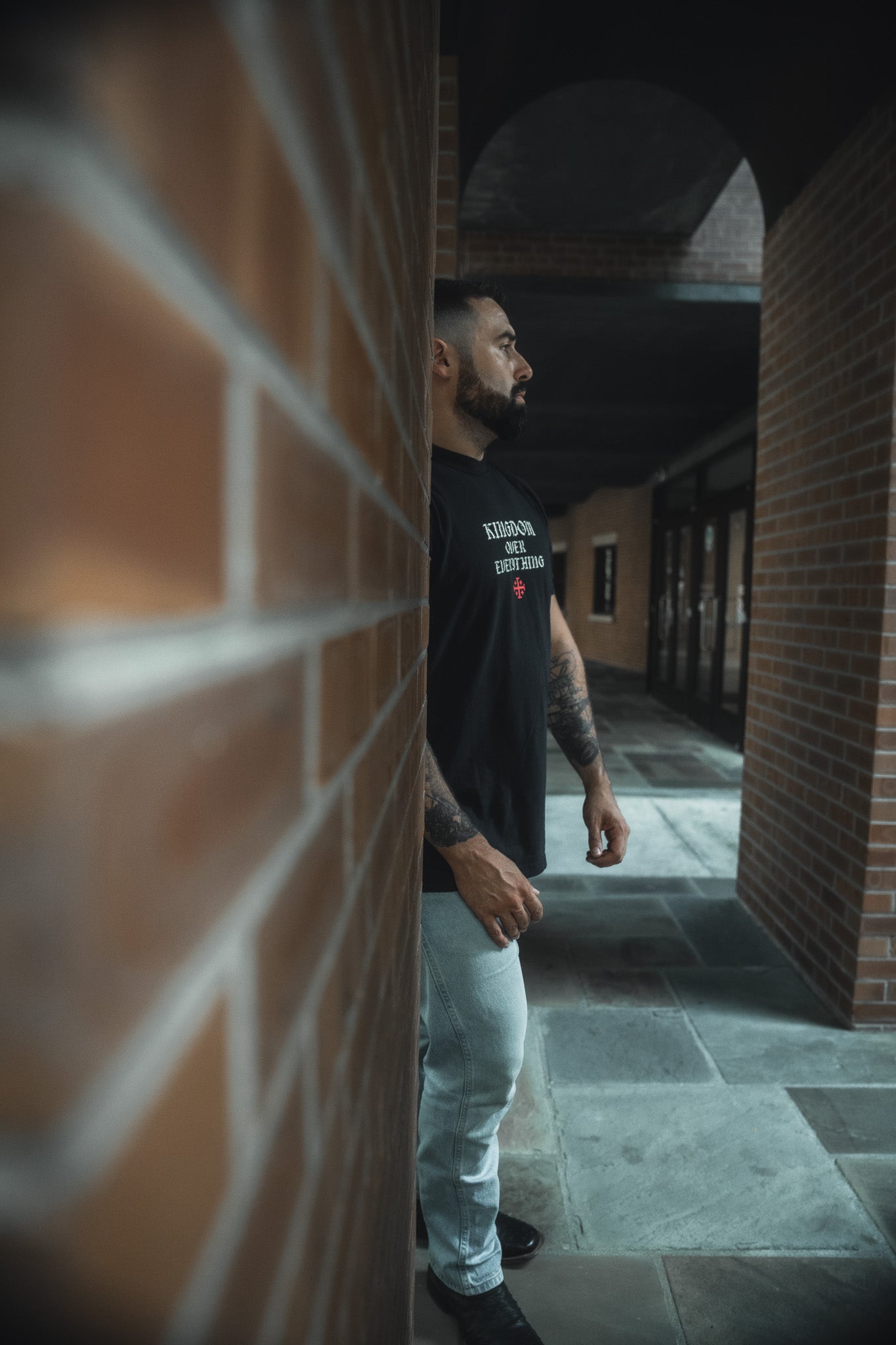 A bearded, tattooed man stands sideways in a brick corridor, wearing the 13th Kingdom Over Everything Premium Tee and light jeans. Soft natural light highlights him as he reflects on seeking His righteousness.