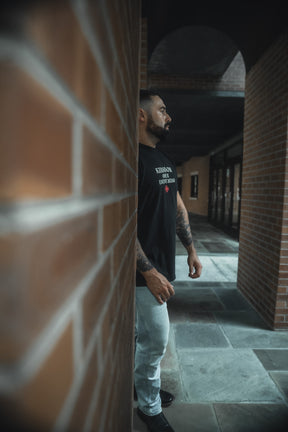A bearded, tattooed man stands sideways in a brick corridor, wearing the 13th Kingdom Over Everything Premium Tee and light jeans. Soft natural light highlights him as he reflects on seeking His righteousness.