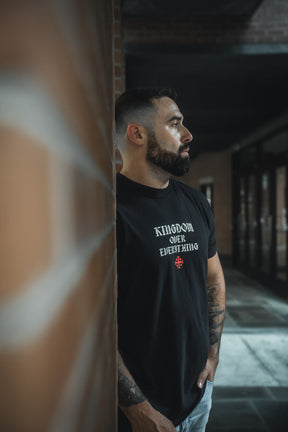 A tattooed man leans against a brick wall, wearing the 13th Kingdom Over Everything Premium Tee featuring a bold KINGDOM OVER EVERYTHING print with a red cross—a reminder of Matthew 6:33 to seek God’s Kingdom first.