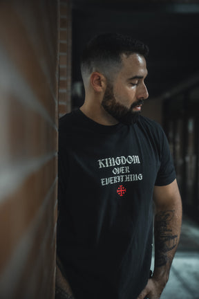 A bearded, tattooed man stands by a brick wall, wearing the 13th Kingdom Over Everything Premium Tee. The black shirt features a red cross and the phrase "KINGDOM OVER EVERYTHING," inspired by Matthew 6:33.