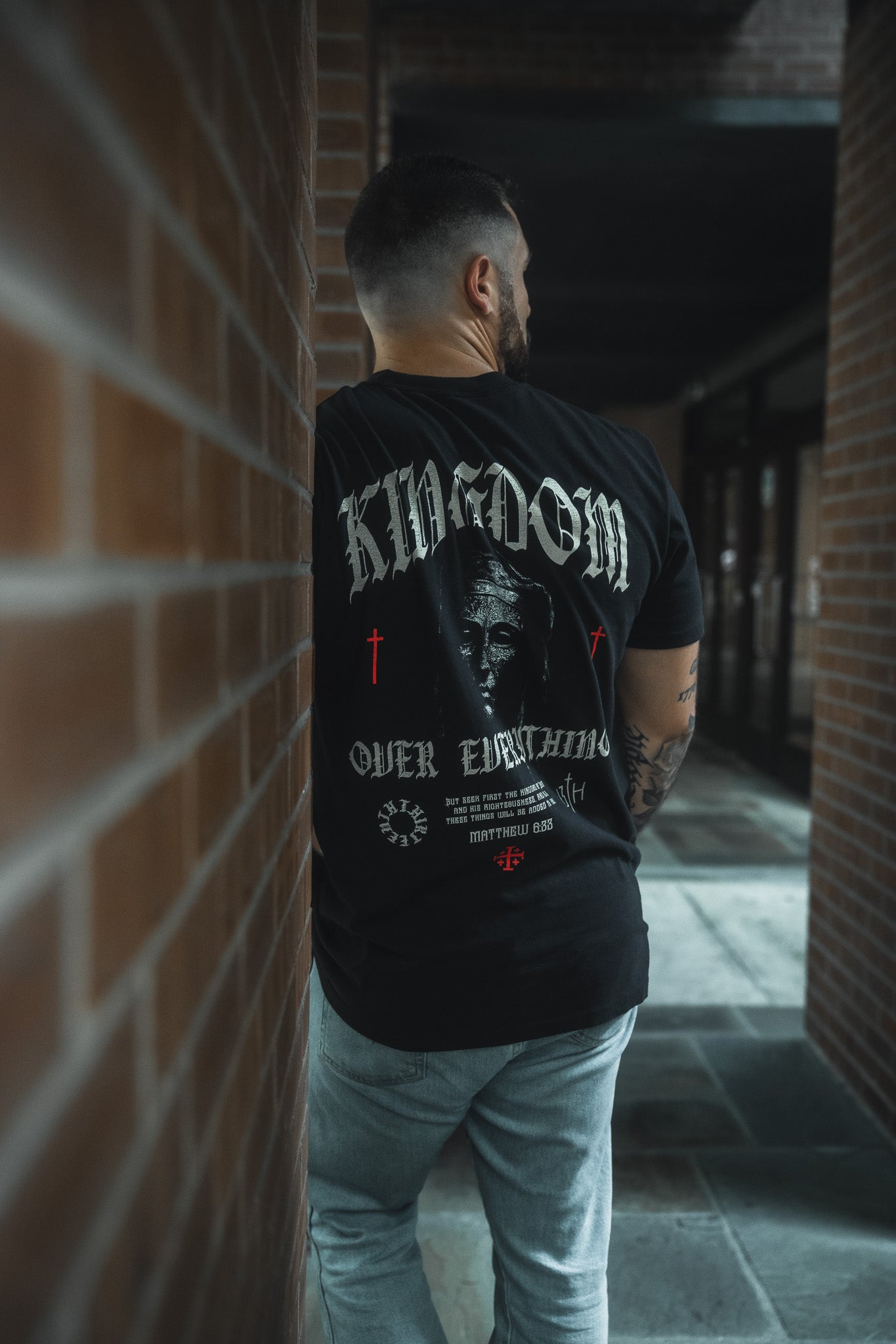 A man with short hair and a trimmed beard stands outdoors between brick walls, facing away. He wears the 13th Kingdom Over Everything Premium Tee with light jeans, bringing spiritual style to the urban, shaded scene.