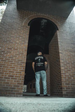 A man in jeans and the 13th Kingdom Over Everything Premium Tee stands under a brick archway, facing away from the camera. The moody urban backdrop echoes the call to seek His righteousness from Matthew 6:33.