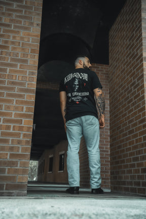 A tattooed man stands in an outdoor corridor of a brick building, back to the camera, wearing the 13th Kingdom Over Everything Premium Tee, which displays "Matthew 6:33" as a reminder to seek first God's kingdom and righteousness.