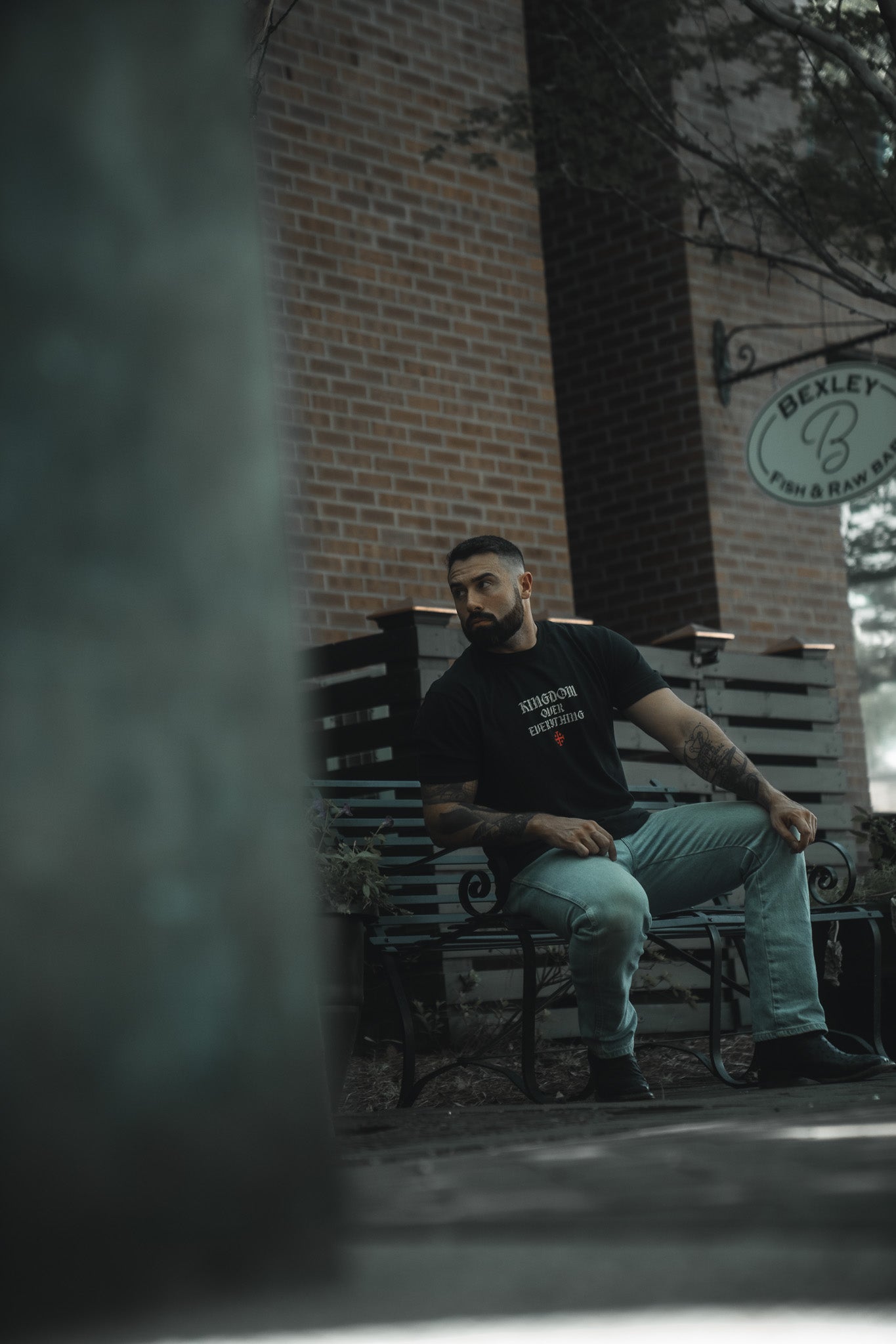 A bearded, tattooed man sits on a black bench outside a brick building with a Bexley Fish & Raw sign, wearing the Kingdom Over Everything Premium Tee by 13th and light blue jeans, looking left as if reflecting on Matthew 6:33.
