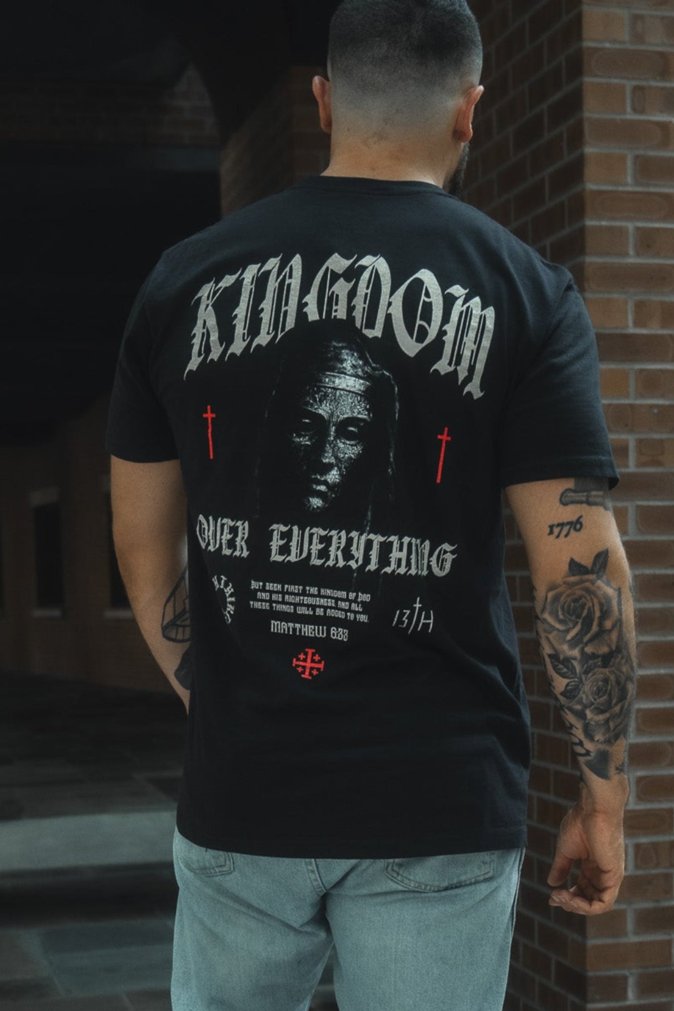 Person wearing a black t-shirt with 'Kingdom Over Everything' text and graphics, standing in front of a brick wall.