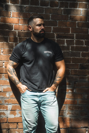 A bearded, tattooed man poses against a brick wall in sunlight, wearing the 13th brand’s The Ten Commandments Premium Tee and light blue jeans—a modern look inspired by timeless Christian values.