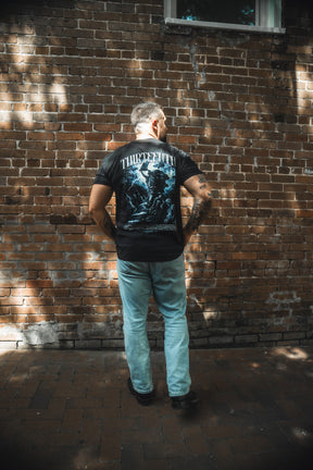 A man with short hair and tattoos stands by a brick wall in light blue jeans and the 13th Ten Commandments Premium Tee, featuring a bold graphic and the word THIRTEENTH on the back. Sunlight dapples the wall behind him.