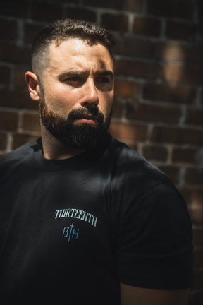A man with short dark hair and a beard stands against a sunlit brick wall, wearing the 13th brand's "The Ten Commandments Premium Tee." He looks off to the side with a serious expression.