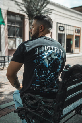 A bearded man sits on a black metal bench outdoors, wearing the 13th brand’s Ten Commandments Premium Tee with bold graphics and “THIRTEENTH” on the back, referencing biblical commandments in a sunlit urban shopping area.