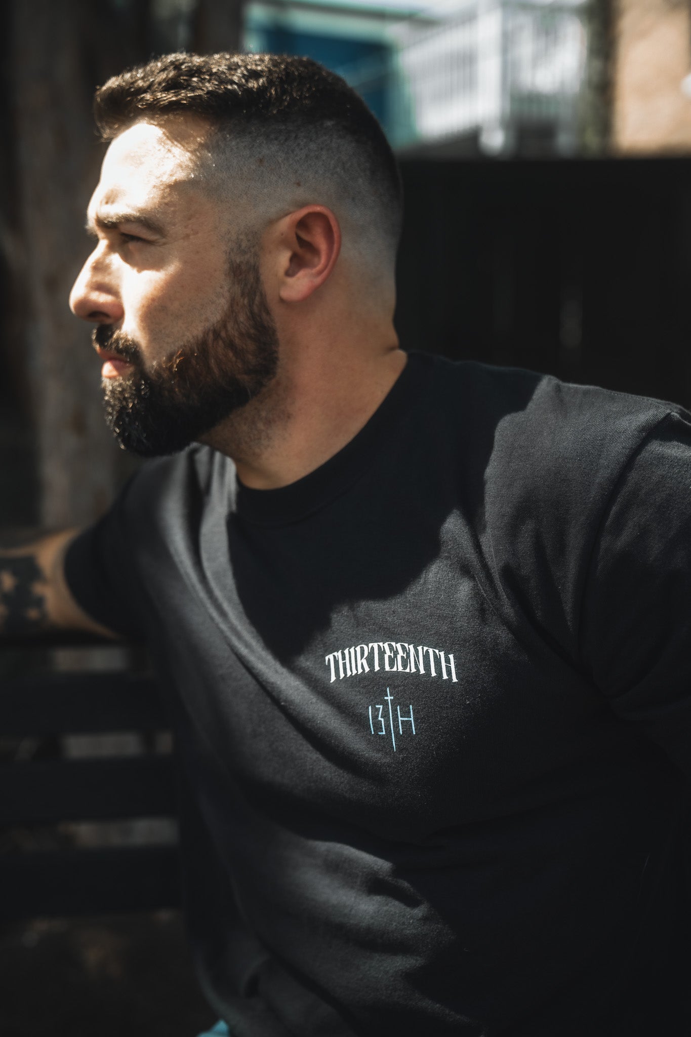 A bearded man with short hair sits outdoors on a bench, wearing the 13th brand's The Ten Commandments Premium Tee, featuring "THIRTEENTH 13TH" in white and blue letters. Sunlight highlights his face as he looks left, evoking Christian values.