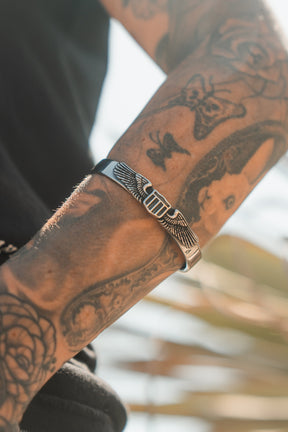 A tattooed arm wearing the Relentless Betrayal Americana Bracelet—a silver, stainless steel piece featuring an engraved wing design and proudly made in the USA—shown in natural sunlight.