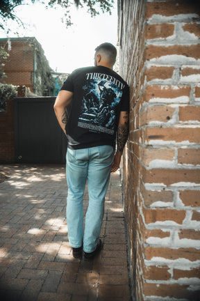 A man stands outdoors near a brick wall, his back to the camera, wearing light blue jeans and the 13th brand’s The Ten Commandments Premium Tee—a black graphic T-shirt.