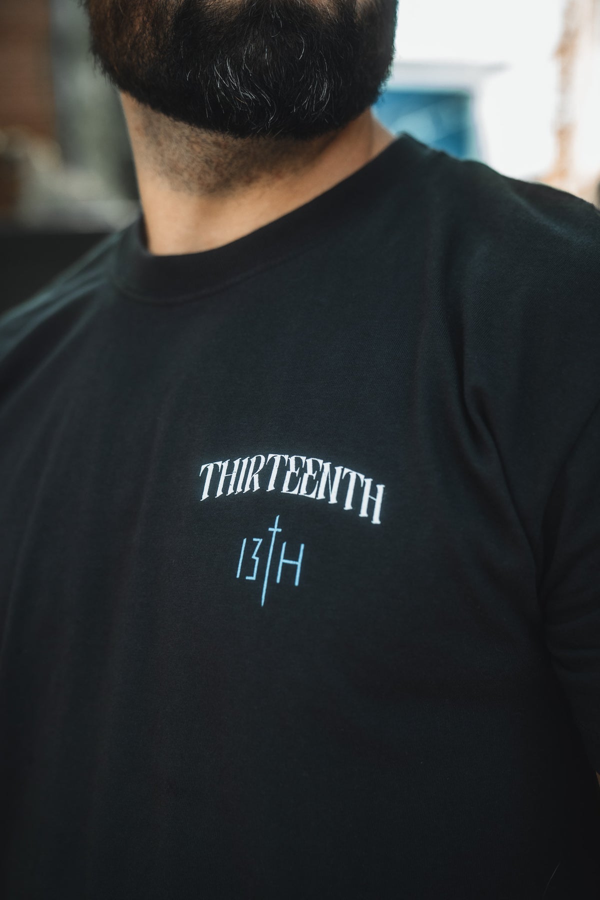 A close-up shows a man wearing the 13th brand's The Ten Commandments Premium Tee, featuring white "THIRTEENTH" text above the number 13 and letter H separated by a vertical line; only his lower face and upper torso are visible.
