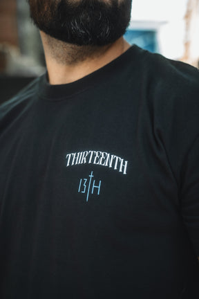 A close-up shows a man wearing the 13th brand's The Ten Commandments Premium Tee, featuring white "THIRTEENTH" text above the number 13 and letter H separated by a vertical line; only his lower face and upper torso are visible.
