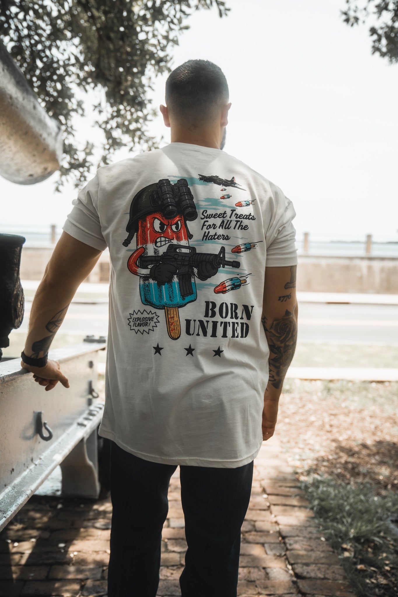 A man stands outdoors facing away, wearing the Born United Private Label "Sweet Victory" white T-shirt with a cartoon popsicle, rockets, and “Sweet Treats For All The Haters” text. Trees and a street are visible in the background.