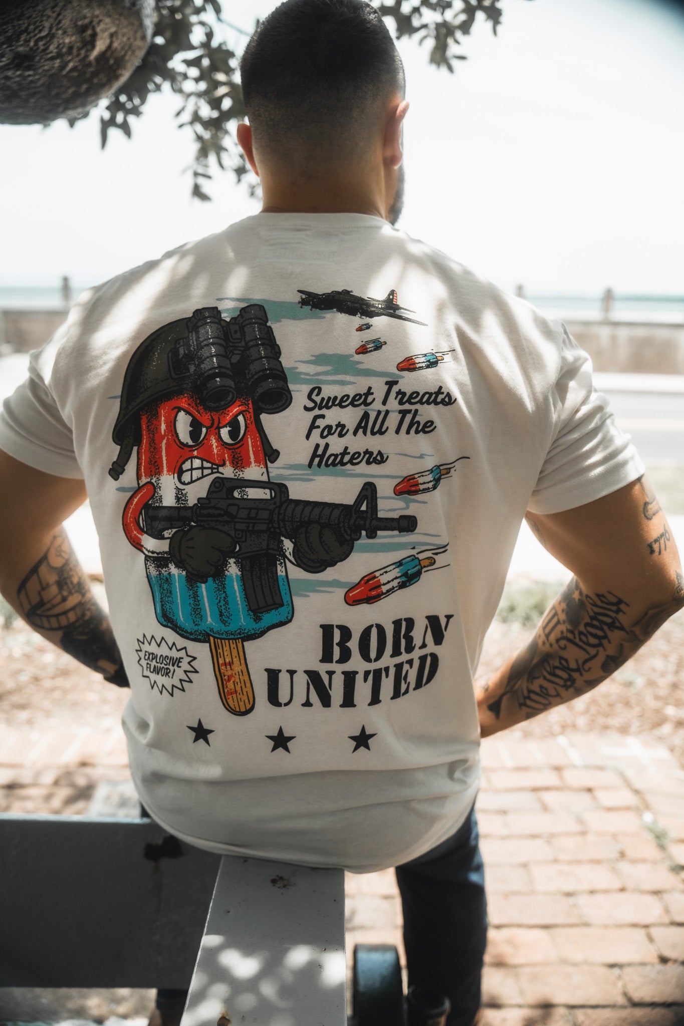 A man sits on an outdoor bench, seen from behind, wearing a Born United Private Label "Sweet Victory" shirt. The shirt shows a cartoon popsicle with helmet and rifle plus the phrases “Sweet Treats For All The Haters” and “BORN UNITED.”.