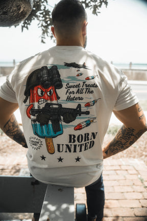 A man sits on an outdoor bench, seen from behind, wearing a Born United Private Label "Sweet Victory" shirt. The shirt shows a cartoon popsicle with helmet and rifle plus the phrases “Sweet Treats For All The Haters” and “BORN UNITED.”.