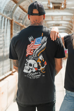 A tattooed man in a black cap and Revolution Premium Tee by Relentless Betrayal stands on a bridge. The shirt’s back shows an eagle with an American flag, skull, and arrows, plus the script “Relentless Betrayal.”.