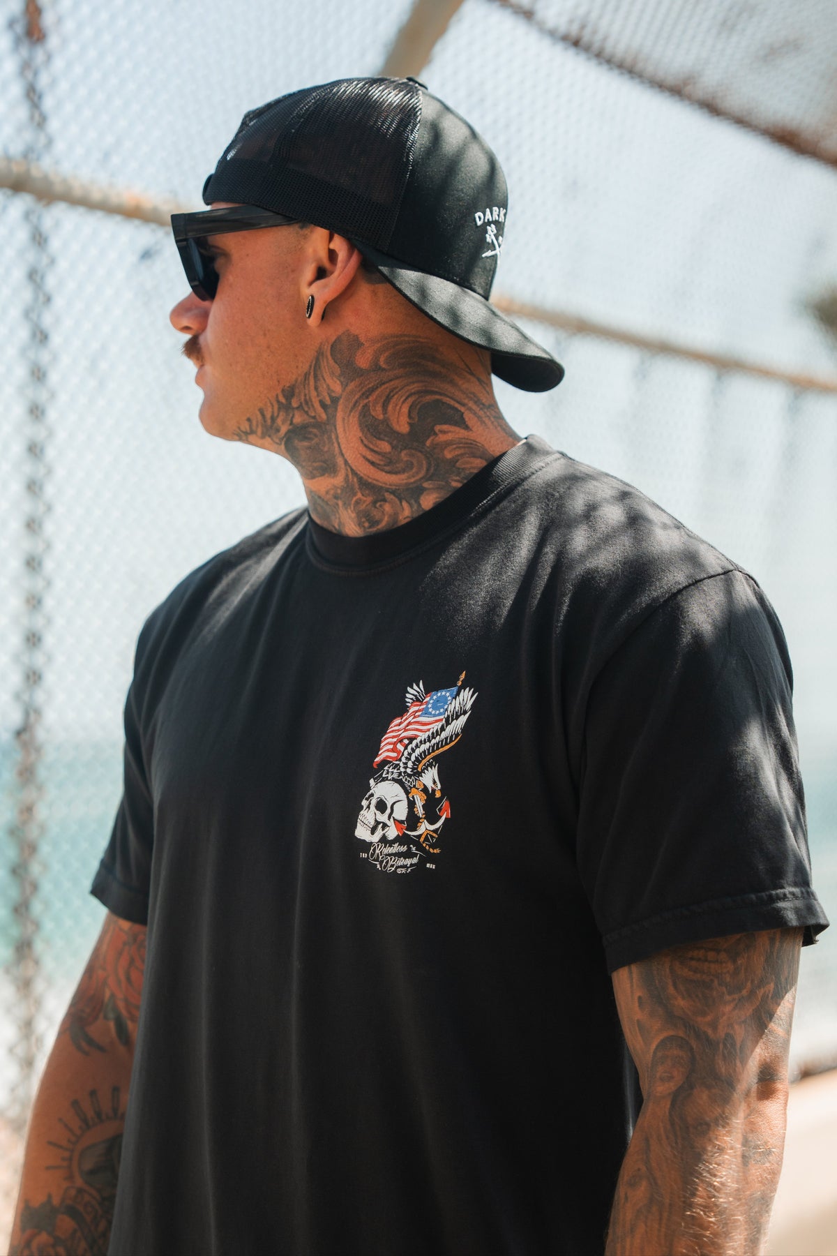 A tattooed person in sunglasses, a backward black cap, and the Relentless Betrayal Revolution Premium Tee featuring an eagle, skull, and American flag design stands outdoors by a wire fence, looking to the side.