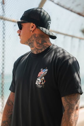 A tattooed person in sunglasses, a backward black cap, and the Relentless Betrayal Revolution Premium Tee featuring an eagle, skull, and American flag design stands outdoors by a wire fence, looking to the side.