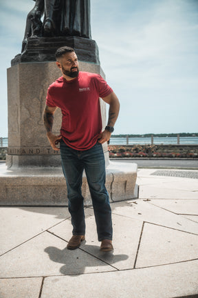 Man in a red t-shirt standing in front of a statue with water and sky in the background